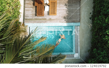 A young woman in a hat and swimsuit is relaxing in the clear water of a private pool 124019792