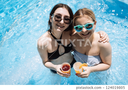 Two smiling girls in swimsuits holding cocktails in the pool on vacation. Cute teenagers in sunglasses are laughing and drinking fresh cool juice. 124019805