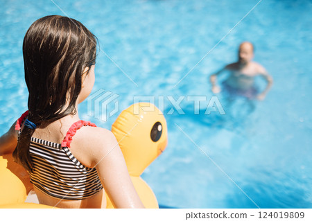 Little girl in swimsuit with inflatable circle is preparing to jump into pool. View from back. Dad and daughter are swimming in blue water. Little girl in swimsuit with inflatable circle is preparing to jump into pool. View from back. Dad and daughter are swimming in blue water. 124019809