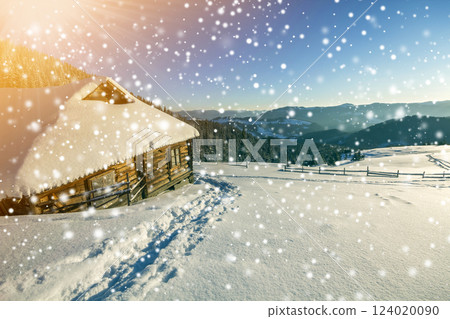 Winter Christmas landscape. Human footprint path in white deep snow at small wooden shepherd hut, spruce forest, woody dark mountain range, large snowflakes on clear blue sky copy space background. 124020090