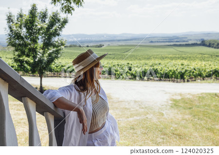 Confident beautiful woman with straw hat and light clothes looking at fields with grape bushes. 124020325