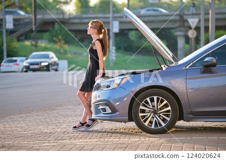 Sad female driver standing on a city street near her car with popped up hood and broken engine waiting for road service assistance to arrive 124020624