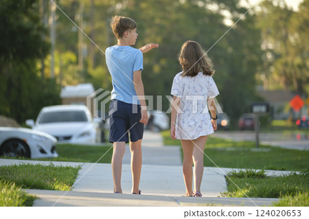Rear view of two young teenage children, girl and boy, brother and sister walking together on rural street on bright sunny day. Vacation time concept Rear view of two young teenage children, girl and boy, brother and sister walking together on rural street on bright sunny day. Vacation time concept 124020653