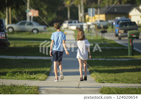 Rear view of two young teenage children, girl and boy, brother and sister walking together on rural street on bright sunny day. Vacation time concept Rear view of two young teenage children, girl and boy, brother and sister walking together on rural street on bright sunny day. Vacation time concept 124020654