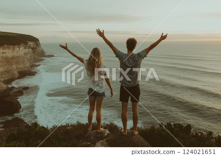 a couple standing on a cliff edge overlooking the ocean with arms raised 124021651
