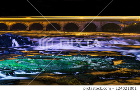 Flowing river under illuminated bridge at night with vibrant reflections and smooth water textures 124021801