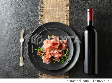 Bottle of red wine and prosciutto on a black plate. 124022323