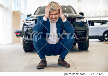 Worried man sitting at his damaged luxury vehicle in workshop 124023250
