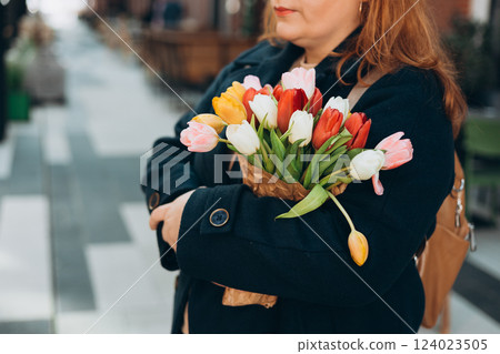 Beautiful young woman with spring tulips flowers bouquet walking at city street. Happy girl posing outdoors. Spring portrait of pretty female on urban background. High quality photo. Beautiful young woman with spring tulips flowers bouquet walking at city street. Happy girl posing outdoors. Spring portrait of pretty female on urban background. High quality photo. 124023505