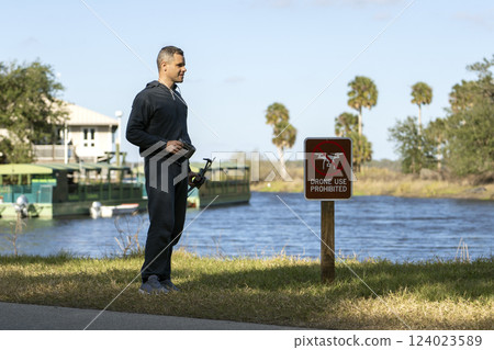 Operator is preparing to fly his quadcopter in national park no drone area. Man unlawfully using his UAV near restriction notice sign Operator is preparing to fly his quadcopter in national park no drone area. Man unlawfully using his UAV near restriction notice sign 124023589
