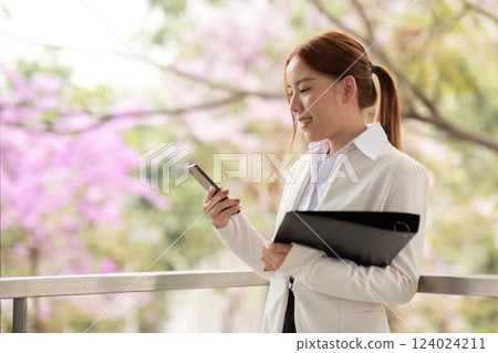 Engaged businesswoman using smartphone while holding files against a backdrop of blooming flowers Engaged businesswoman using smartphone while holding files against a backdrop of blooming flowers 124024211