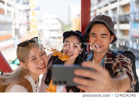Young Asian friends taking a joyful selfie while enjoying street food at a lively market, celebrating travel and friendship. 124024240