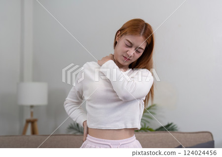 Young woman grimacing and holding her shoulder and back, seeking comfort from pain in her living space Young woman grimacing and holding her shoulder and back, seeking comfort from pain in her living space 124024456