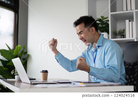Joyful Asian businessman celebrating success at his desk, raising fists in excitement while working on a laptop in a modern office. 124024516