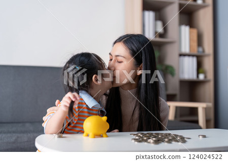 Diverse mother sharing a loving kiss with her daughter while saving money with a yellow piggy bank and coins at home, promoting family bonding. 124024522