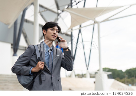 Cheerful young Asian businessman talking on smartphone while carrying a bag outdoors, dressed in professional suit 124024526
