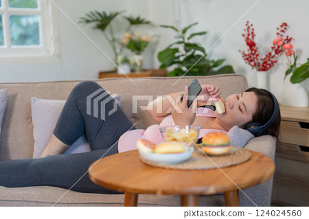 Overweight woman relaxing on the couch, enjoying snacks while scrolling on her phone at home 124024560