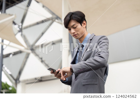 Young Asian businessman checking watch while using smartphone outdoors, dressed in formal suit 124024562