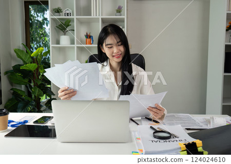 Financial Document Evaluation. An individual assessing tax documents with a smile at a workspace filled with paperwork and a laptop. Financial Document Evaluation. An individual assessing tax documents with a smile at a workspace filled with paperwork and a laptop. 124024610