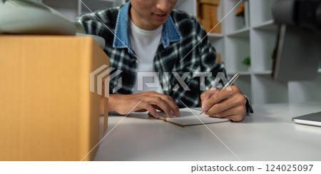 Young entrepreneur taking notes for online sales strategy, writing in notebook at modern workspace with product boxes. 124025097
