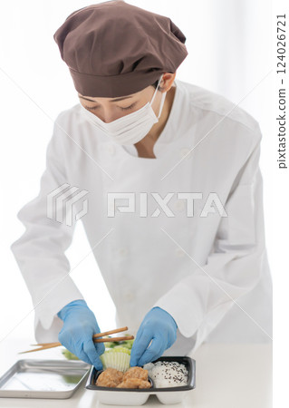 一名戴著口罩的女子將配菜放入便當盒中 一名戴著口罩的女子將配菜放入便當盒中 124026721