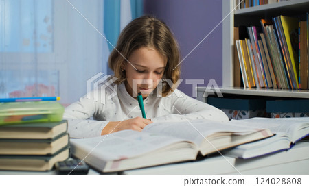 Immersed in her studies, a young girl works at her desk in a warm room filled with books in the evening 124028808
