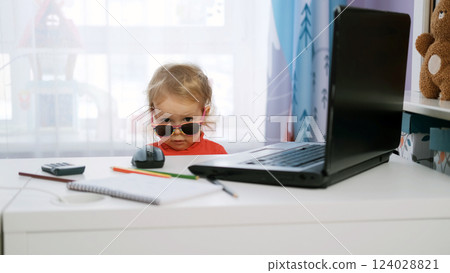 Baby girl adds sunglasses to her look while seated at a desk with a laptop, notebook, and stationery Baby girl adds sunglasses to her look while seated at a desk with a laptop, notebook, and stationery 124028821
