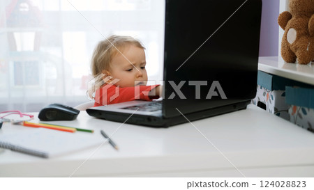 Cute baby girl sits at her desk typing on a laptop, surrounded by coloring pens and a notebook. Young freelancer working in a cozy space 124028823