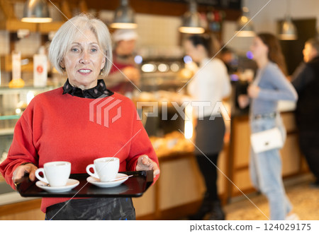 Senior woman visitor in self-service cafeteria carries tray with two cups of coffee. Senior woman visitor in self-service cafeteria carries tray with two cups of coffee. 124029175