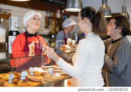 Bakery shop senior female assistant talk to customer, answers questions and recommends fresh products 124029232
