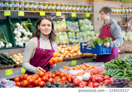 Young female seller holding tomatoes standing by counter in grocery market 124029337