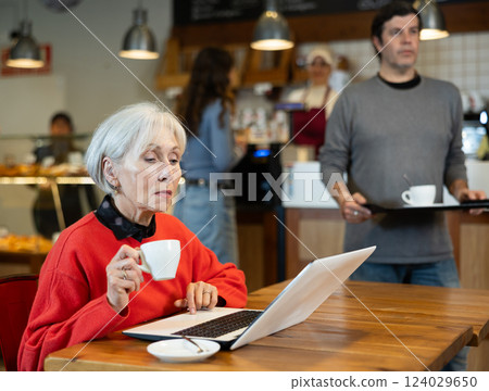 In cozy cafe hall, freelance senior woman works with laptop and drinks coffee. In cozy cafe hall, freelance senior woman works with laptop and drinks coffee. 124029650
