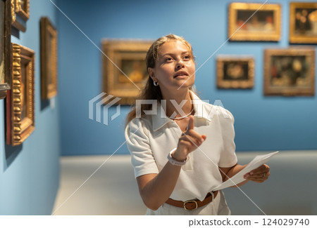 Young girl holding an information booklet looks at paintings in an art exhibition in museum Young girl holding an information booklet looks at paintings in an art exhibition in museum 124029740