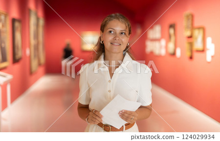 Young girl holding an information booklet looks at paintings in an art exhibition in museum 124029934