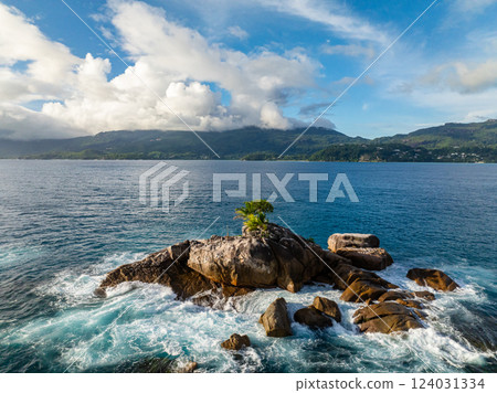 A small rocky island with a tree stands in turquoise water as waves crash around it, surrounded by the ocean. Seychelles, Mahe. 124031334
