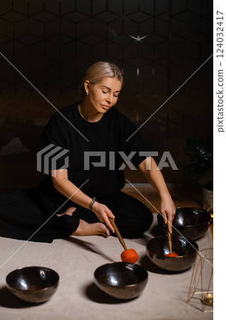 Woman practicing sound healing with singing bowls in a tranquil indoor space decorated with plants and soft lighting during a peaceful evening session 124032417