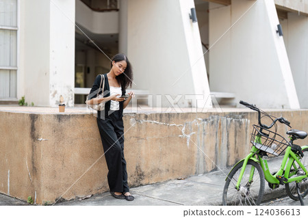 Technology and Convenience. A young woman stands casually, focused on her smartphone while leaning against a wall near her bicycle. Technology and Convenience. A young woman stands casually, focused on her smartphone while leaning against a wall near her bicycle. 124036613
