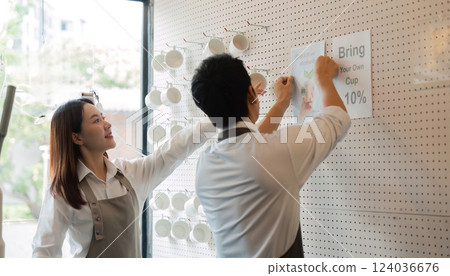 Engagement and Promotion. Two baristas hanging promotional materials for sustainability in their coffee shop, showcasing eco-friendly practices. 124036676
