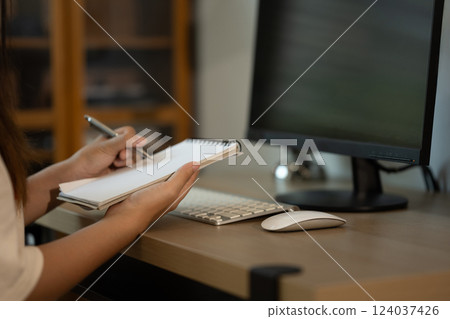 Cropped shot woman is writing in notebook while sitting at a desk with a computer monitor Cropped shot woman is writing in notebook while sitting at a desk with a computer monitor 124037426