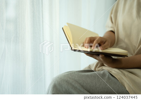 Peaceful young woman holding an open notebook sitting by a softly lit window Peaceful young woman holding an open notebook sitting by a softly lit window 124037445