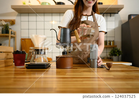 Smiling woman in an apron pouring coffee beans into hand grinder, preparing fresh brew in a home kitchen Smiling woman in an apron pouring coffee beans into hand grinder, preparing fresh brew in a home kitchen 124037525