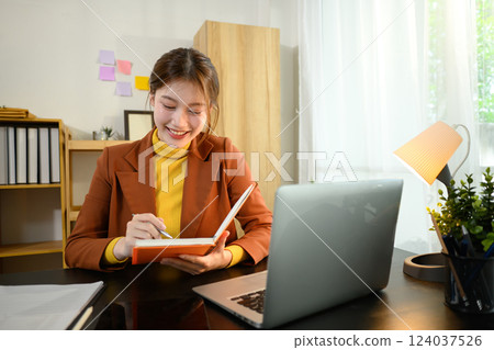 Smiling businesswoman writing in a notebook while working on her laptop at desk 124037526