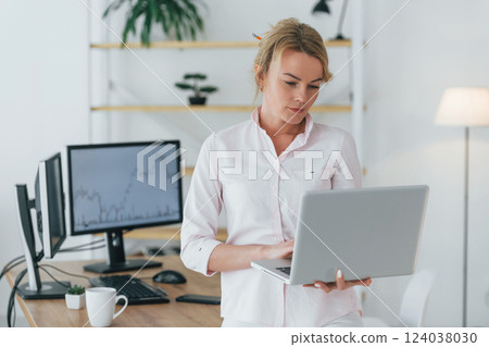 Standing with laptop in hands. Female stock broker in formal wear is working in the office 124038030