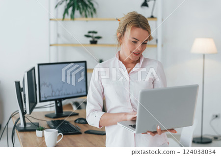 Standing with laptop in hands. Female stock broker in formal wear is working in the office Standing with laptop in hands. Female stock broker in formal wear is working in the office 124038034