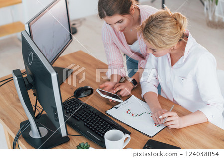 Talking with each other. Two female stock brokers in formal wear is working in the office by pc Talking with each other. Two female stock brokers in formal wear is working in the office by pc 124038054