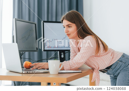Standing with apple in hand and looking at laptop. Female stock broker in casual clothes is working in the office by pc 124038088