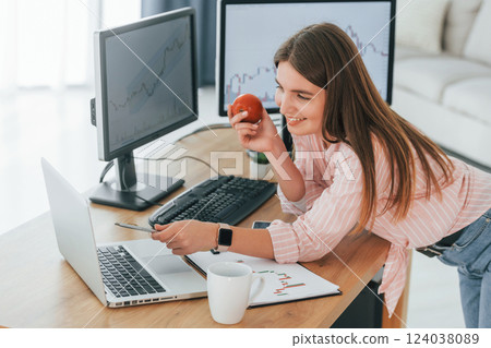 Standing with apple in hand and looking at laptop. Female stock broker in casual clothes is working in the office by pc 124038089