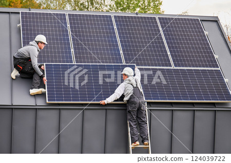 Mounters building photovoltaic solar module station on roof of house. Men electricians in helmets installing solar panel system outdoors. Concept of alternative and renewable energy. 124039722