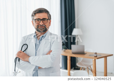 Standing and holding stethoscope. Professional medical worker in white coat is in the office Standing and holding stethoscope. Professional medical worker in white coat is in the office 124040525