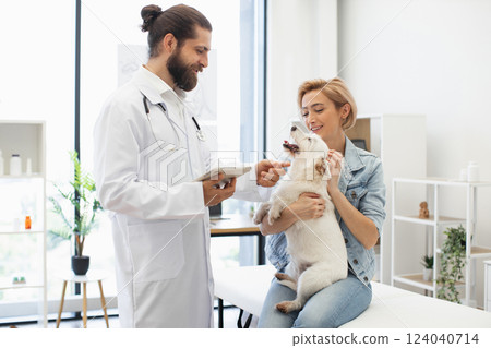 Man veterinarian with young adult woman owner examining white dog at modern clinic. Smiling doctor providing care. Concept of pet health, animal care, veterinary service, friendship. 124040714
